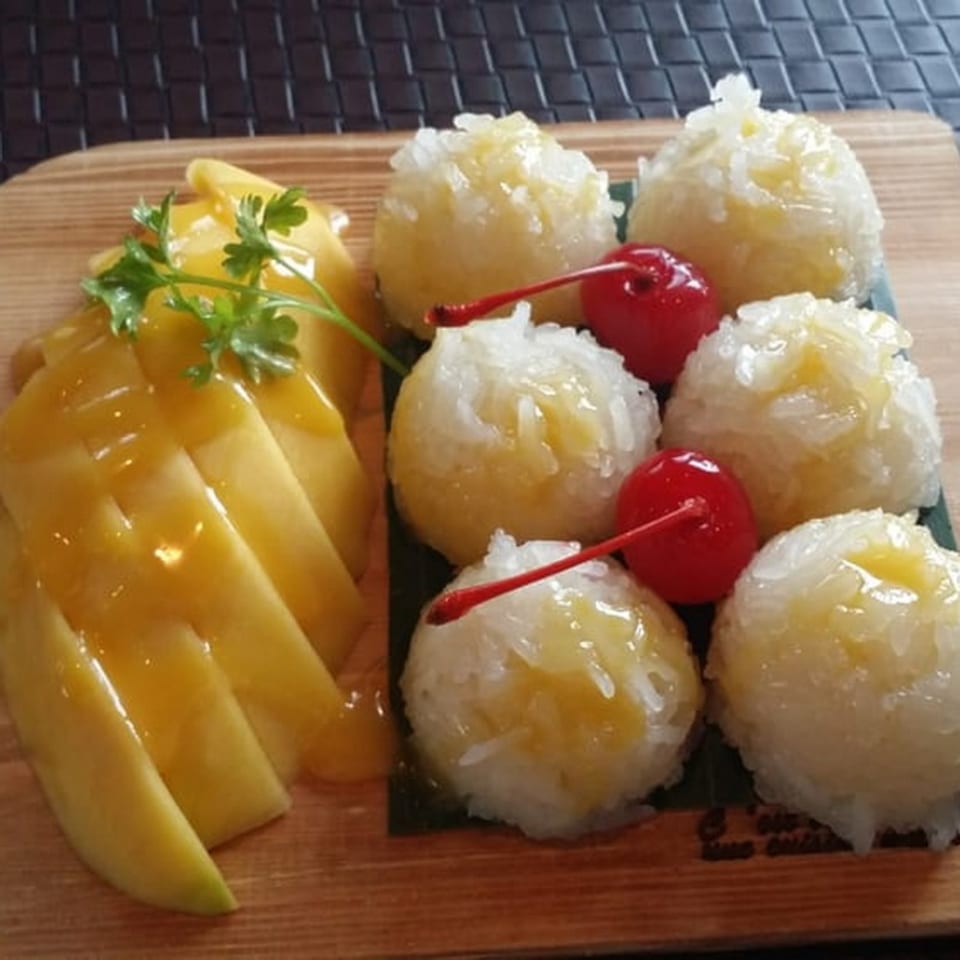 Thai mango sticky rice dessert with sliced mango, sticky rice balls, and cherries on a wooden board.