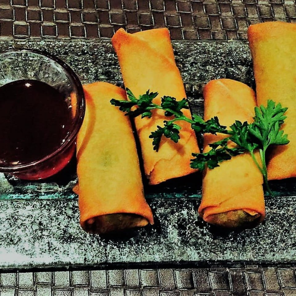 Crispy golden spring rolls garnished with parsley, served with a dark dipping sauce on a textured tray.