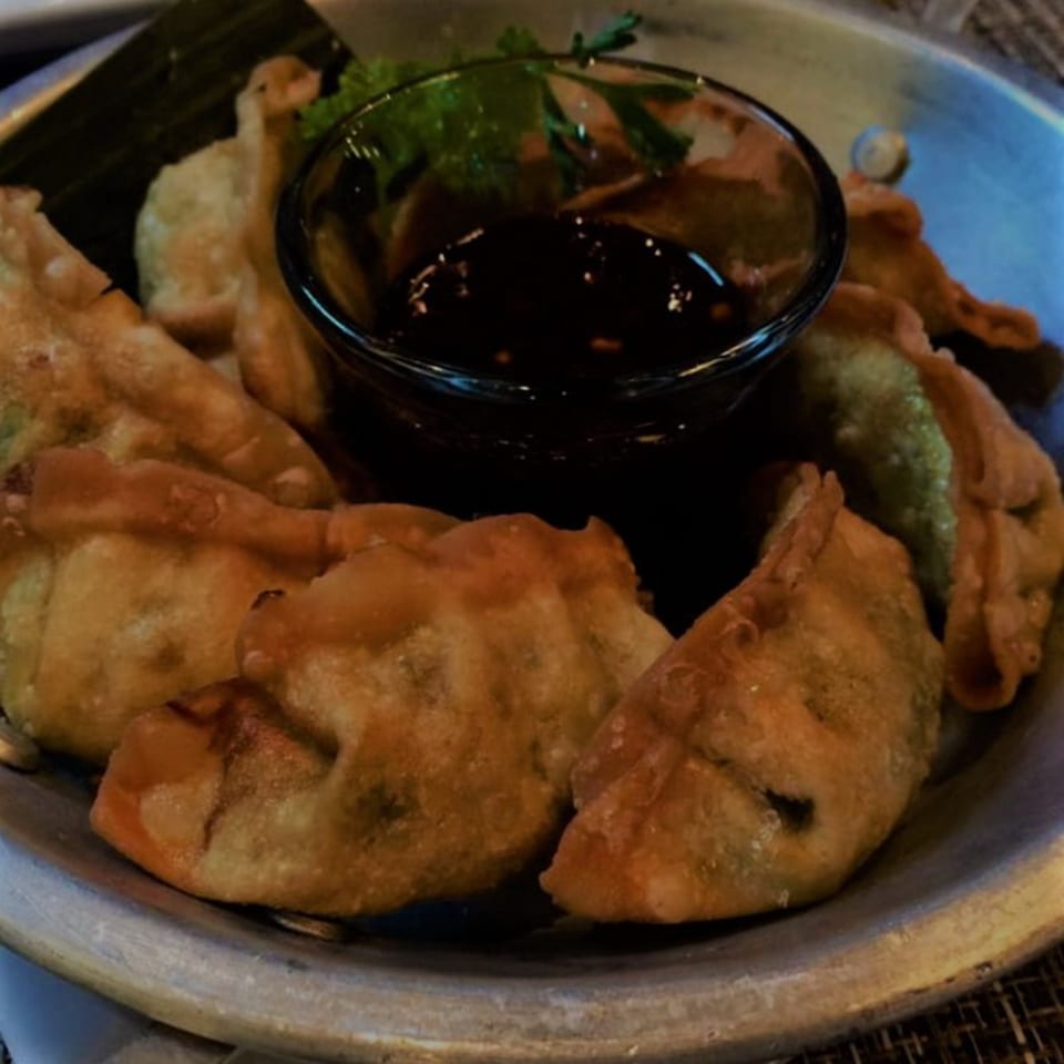 Golden-brown fried Thai dumplings with a side of dark dipping sauce and green garnish in a metal bowl.