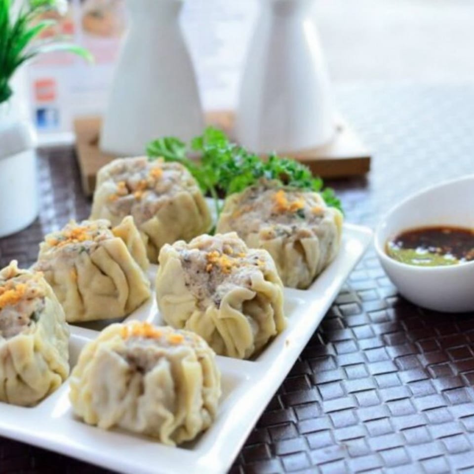 A white rectangular plate holds six Thai dumplings, garnished with herbs and a side of dipping sauce.