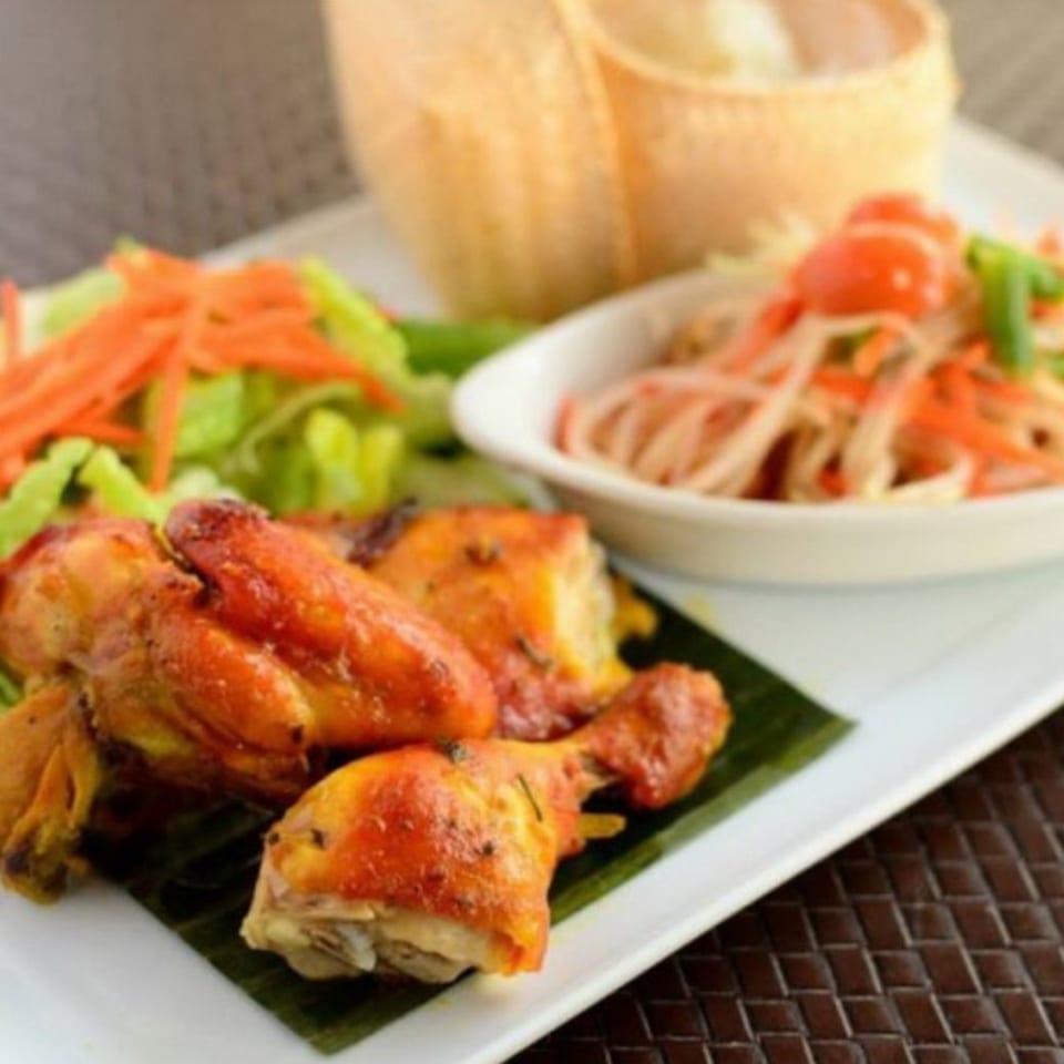 Grilled chicken drumsticks with fresh salad, papaya salad, and sticky rice on a white plate.