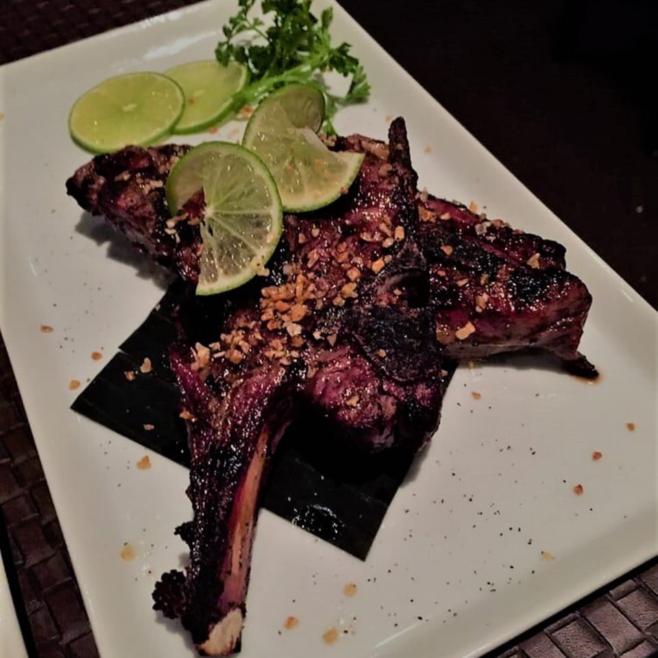 Grilled lamb chops with lime slices and chopped peanuts on a white rectangular plate.