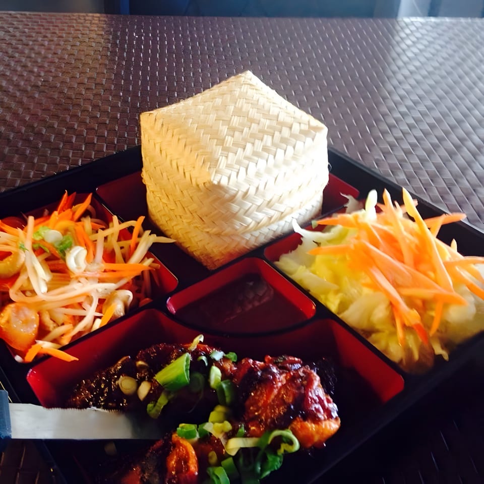 A Thai bento box with chicken, papaya salad, cabbage, and sticky rice in a woven basket.