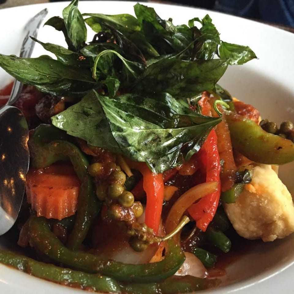 Close-up of a Thai dish with green basil, red peppers, green beans, and carrots in a rich sauce.