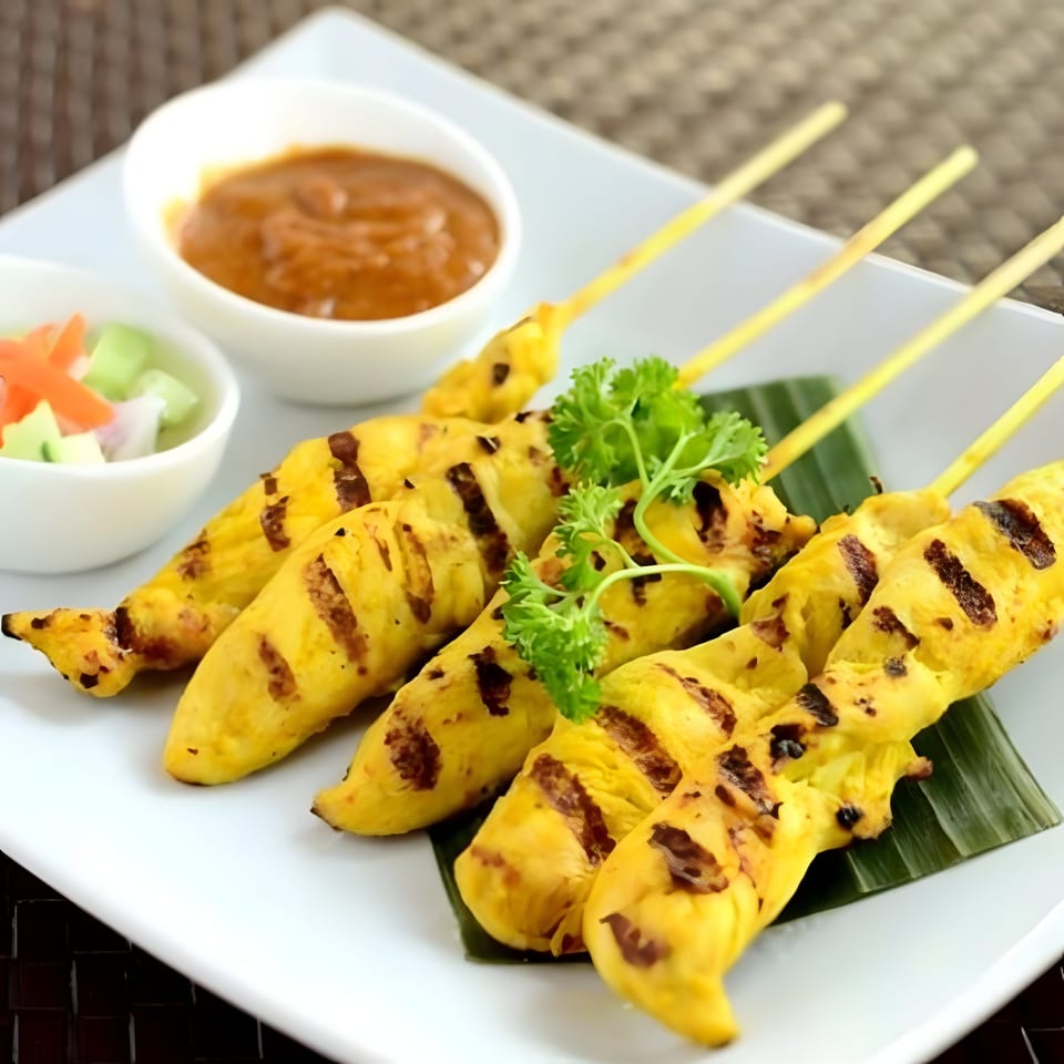 Grilled chicken satay skewers with peanut sauce and cucumber relish on a white plate.