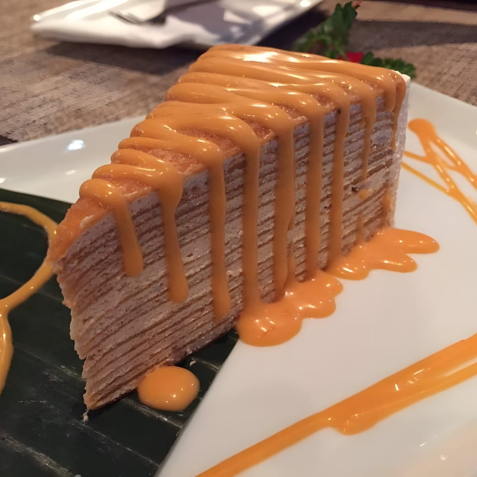 A slice of Thai tea crepe cake with orange drizzle on a white plate with a banana leaf.