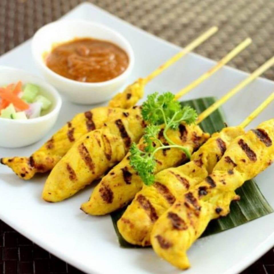 Grilled chicken satay skewers with peanut sauce and cucumber relish on a white plate.